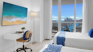 Ein Hotelzimmer mit Blick auf die Stadt, ein Schreibtisch mit Stuhl, ein Bett mit blauer Bettwäsche und ein Strandbild.