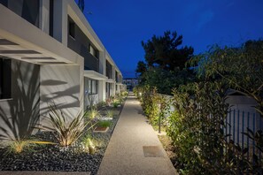 Un camino bordeado por edificios modernos, un jardín bien cuidado con diversas plantas y una cerca azul.
