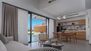 A modern living room with a sofa, a dining area with wicker chairs, a kitchen with marble countertops, and a sliding glass door to the outside. - Viridian Exclusive Villas (Sithonia)