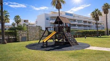 A playground with a slide and climbing area, surrounded by grass and palm trees, with a multi-story building in the background.