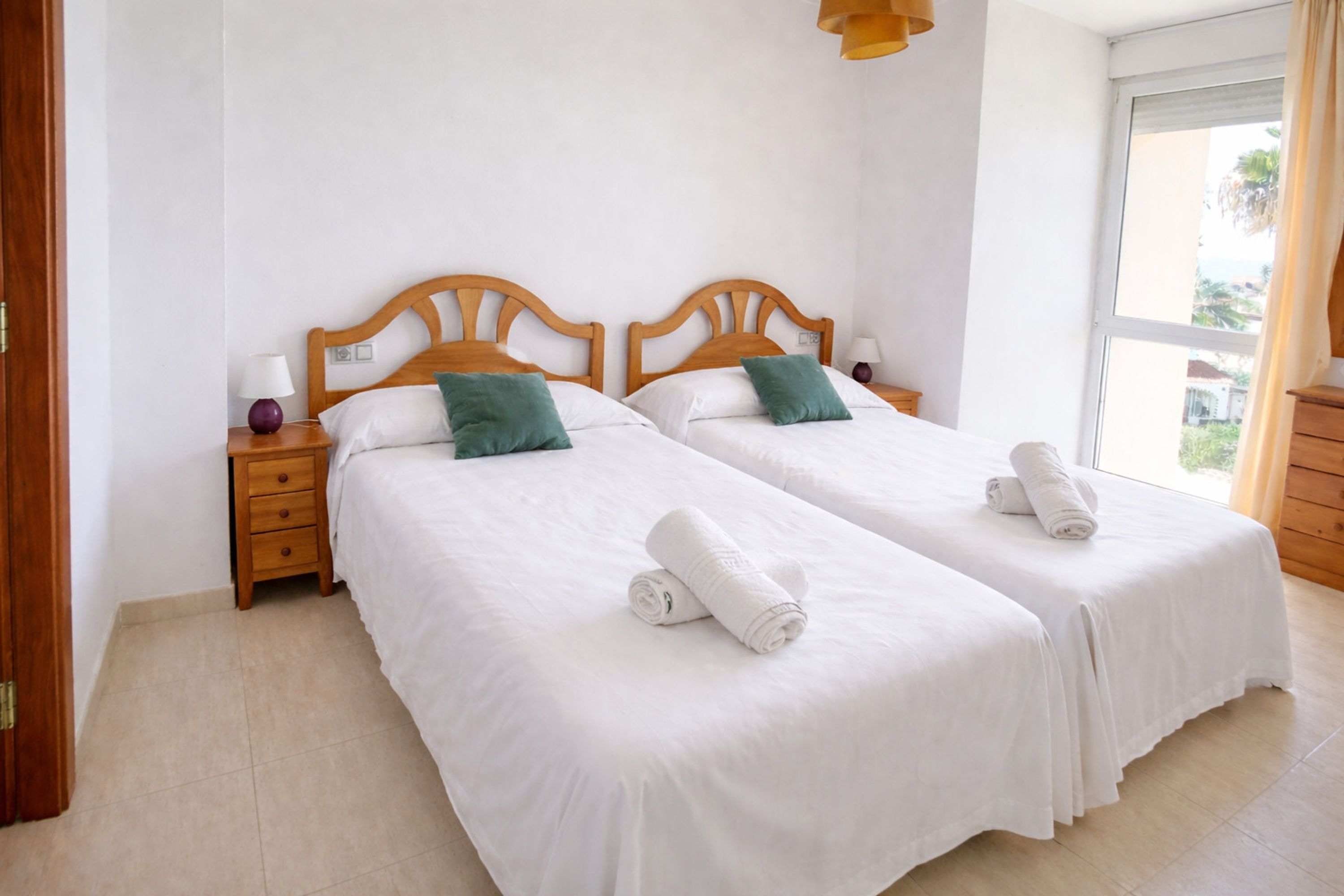 Quarto de hotel com duas camas de solteiro, cabeceiras de madeira, roupa de cama branca e janela com vista.