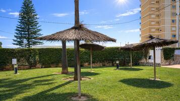 A grassy area with thatched umbrellas, a tall tree, and a building in the background.