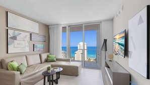 A modern living room with a beige sofa, a small round table, and a large window offering a view of the ocean and buildings. - Ocean Bliss Retreat - 2 BR. Apartment (Hollywood)