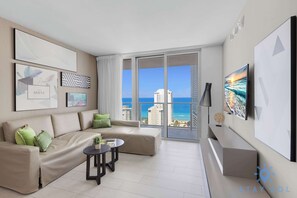 A modern living room with a beige sofa, a small round table, and a large window offering a view of the ocean and buildings. - Ocean Bliss Retreat - 2 BR. Apartment (Hollywood)
