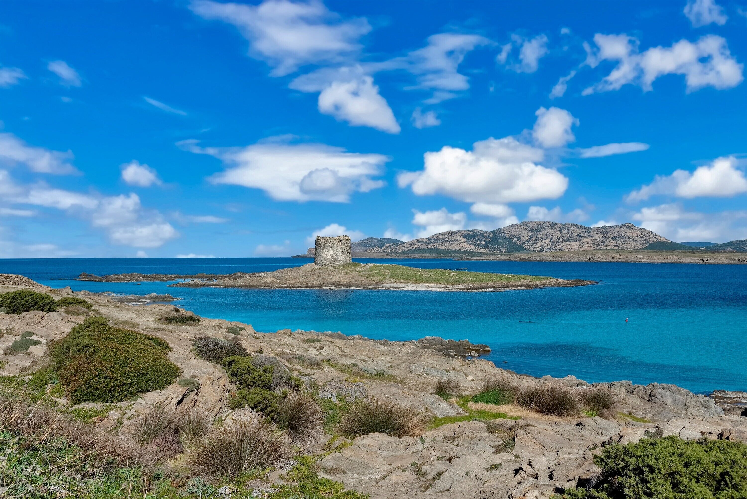 Un paisaje costero con una estructura de piedra histórica, terreno rocoso y aguas azules cristalinas.