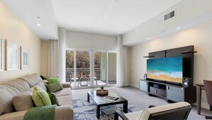 Een moderne woonkamer met een bank, salontafel en een flatscreen televisie. Er is een balkon met uitzicht op bomen en een hekwerk.
