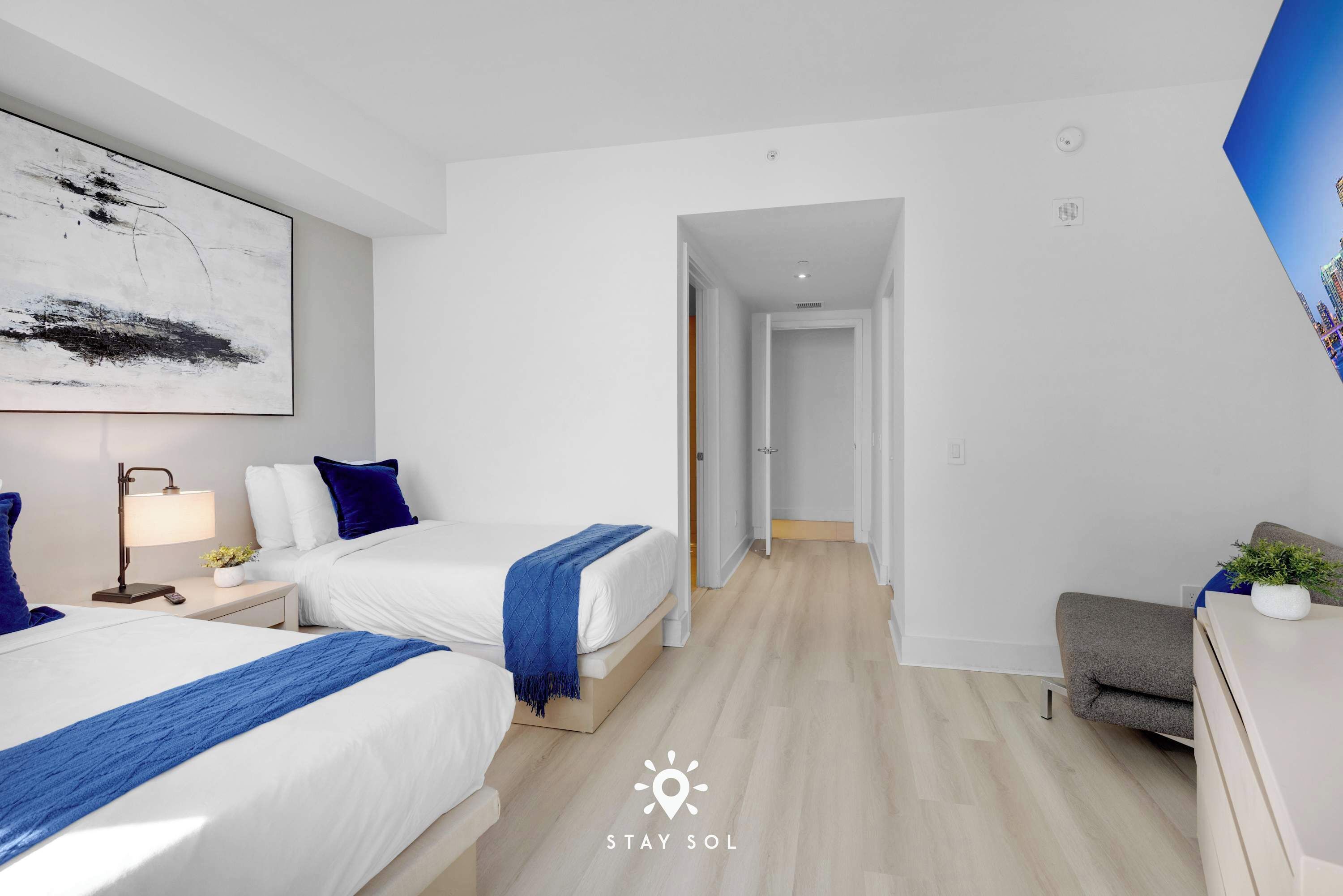 A modern hotel room with two beds, a desk, a chair, and a large abstract painting on the wall.