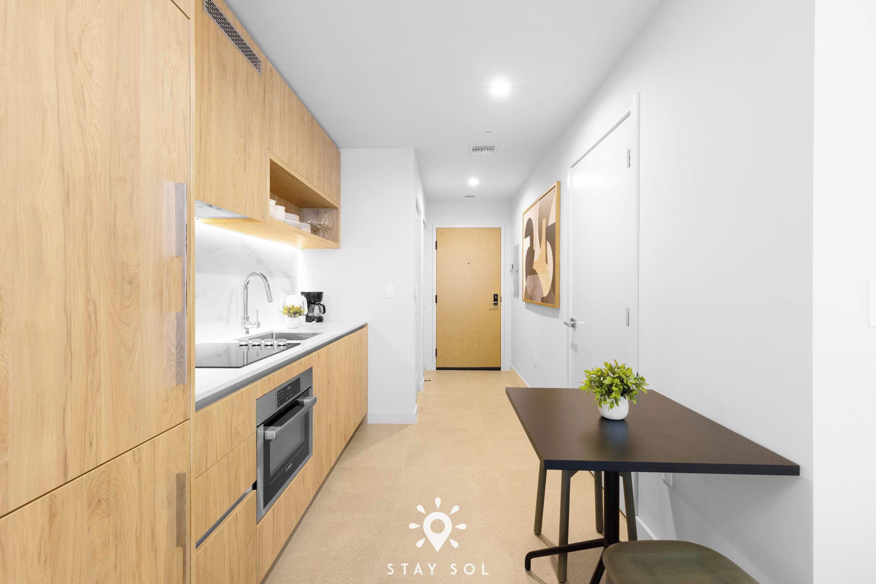 A modern kitchen with wooden cabinets, a built-in oven, and a sink. There is a small table with a plant and a door leading to another room.