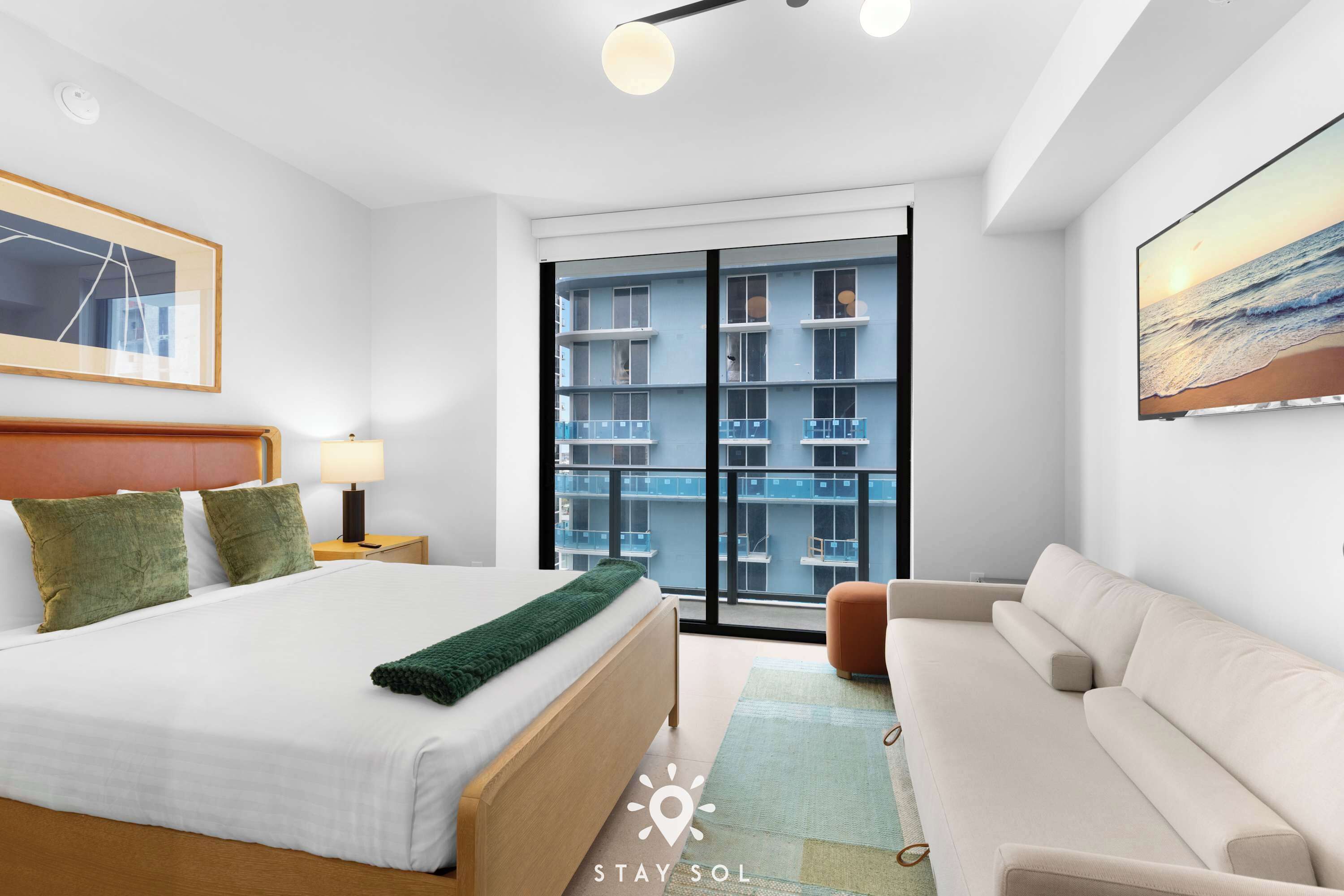A modern hotel room with a large bed, a sofa, a view of a building through a glass door, and a painting on the wall.