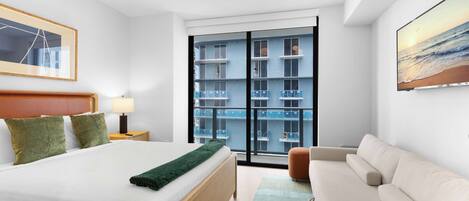 A modern hotel room with a large bed, a sofa, a view of a building through a glass door, and a painting on the wall.