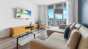 A modern living room with a large window, a flat-screen TV, a sofa, a glass coffee table, and a wooden cabinet. - Cozy Upper Apartment | Bay View Fitness Center (Hallandale Beach)