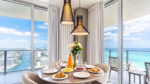 Ein Essbereich mit einem Tisch für vier Personen, einer Blumenvase und Blick auf das Meer sowie die Stadtlandschaft durch große Fenster.