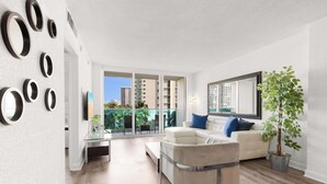 A modern living room with a white sofa, a coffee table, and a large mirror. There is a potted plant and a balcony with a view of buildings. - Modern Hollywood Beach Escape - City Views (Hollywood)