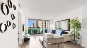 A modern living room with a white sofa, a coffee table, and a large mirror. There is a potted plant and a balcony with a view of buildings. - Modern Hollywood Beach Escape - City Views (Hollywood)