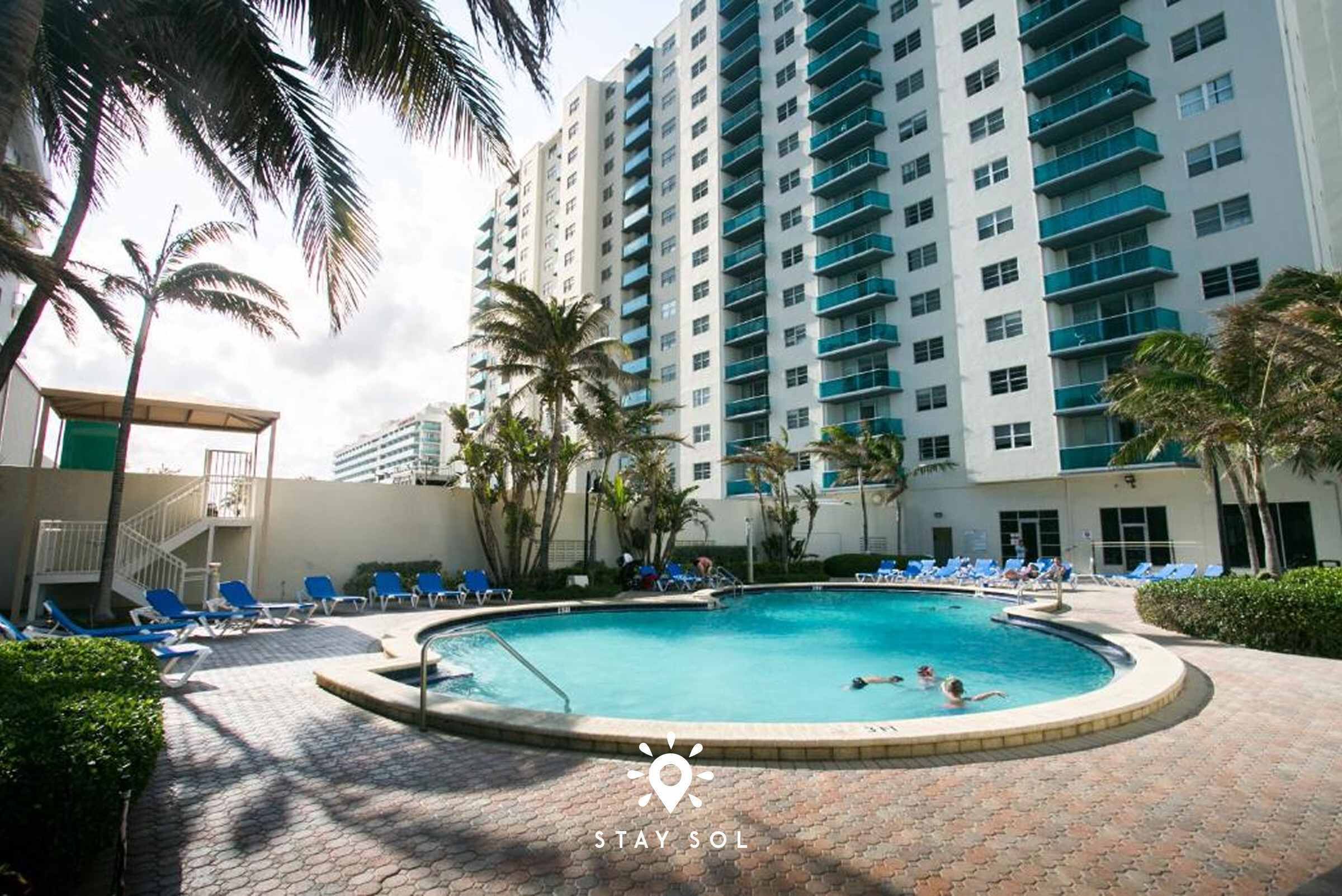 A swimming pool surrounded by blue lounge chairs, palm trees, and a tall building with balconies.