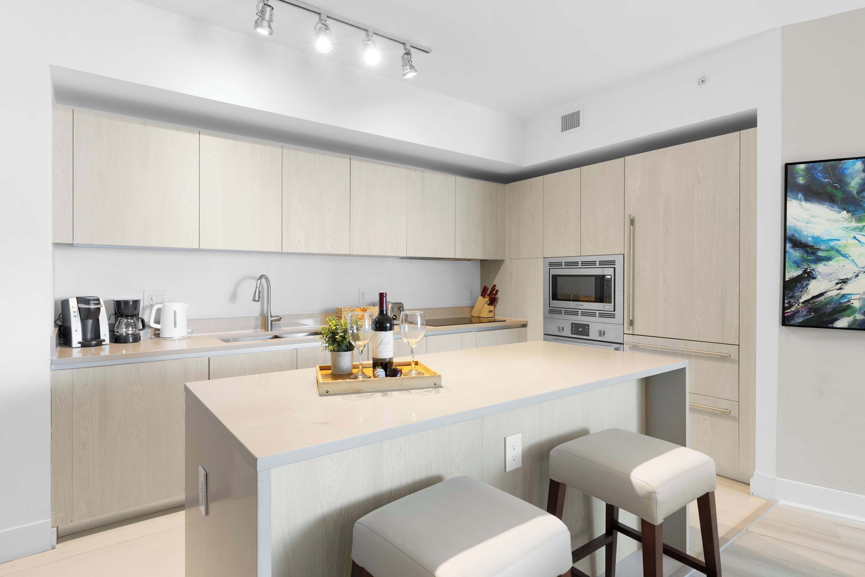 A modern kitchen with light wood cabinets, a central island with bar stools, a built-in oven, and a coffee machine.