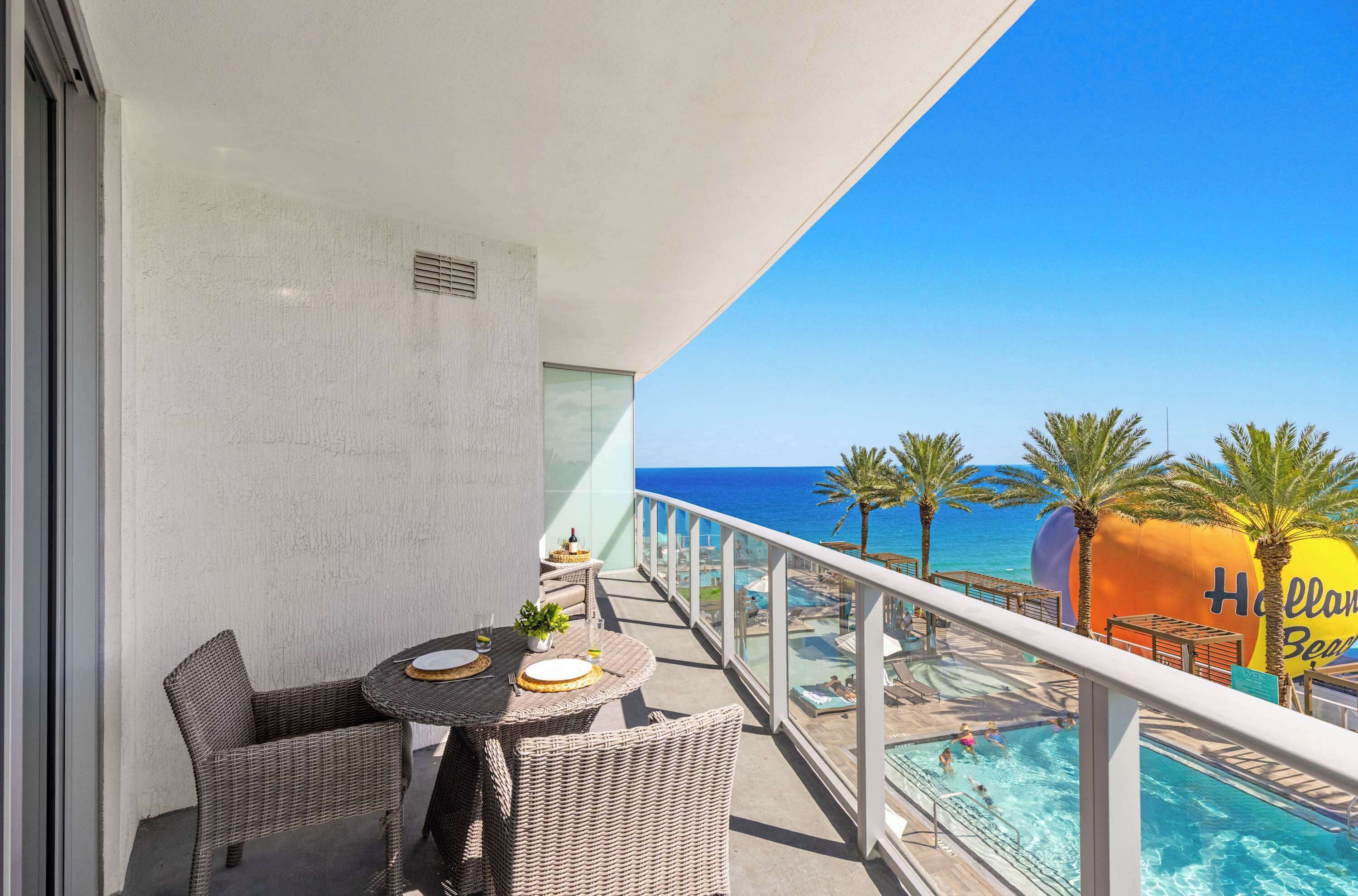 A balcony with wicker furniture, a view of the ocean, and a pool area with palm trees.