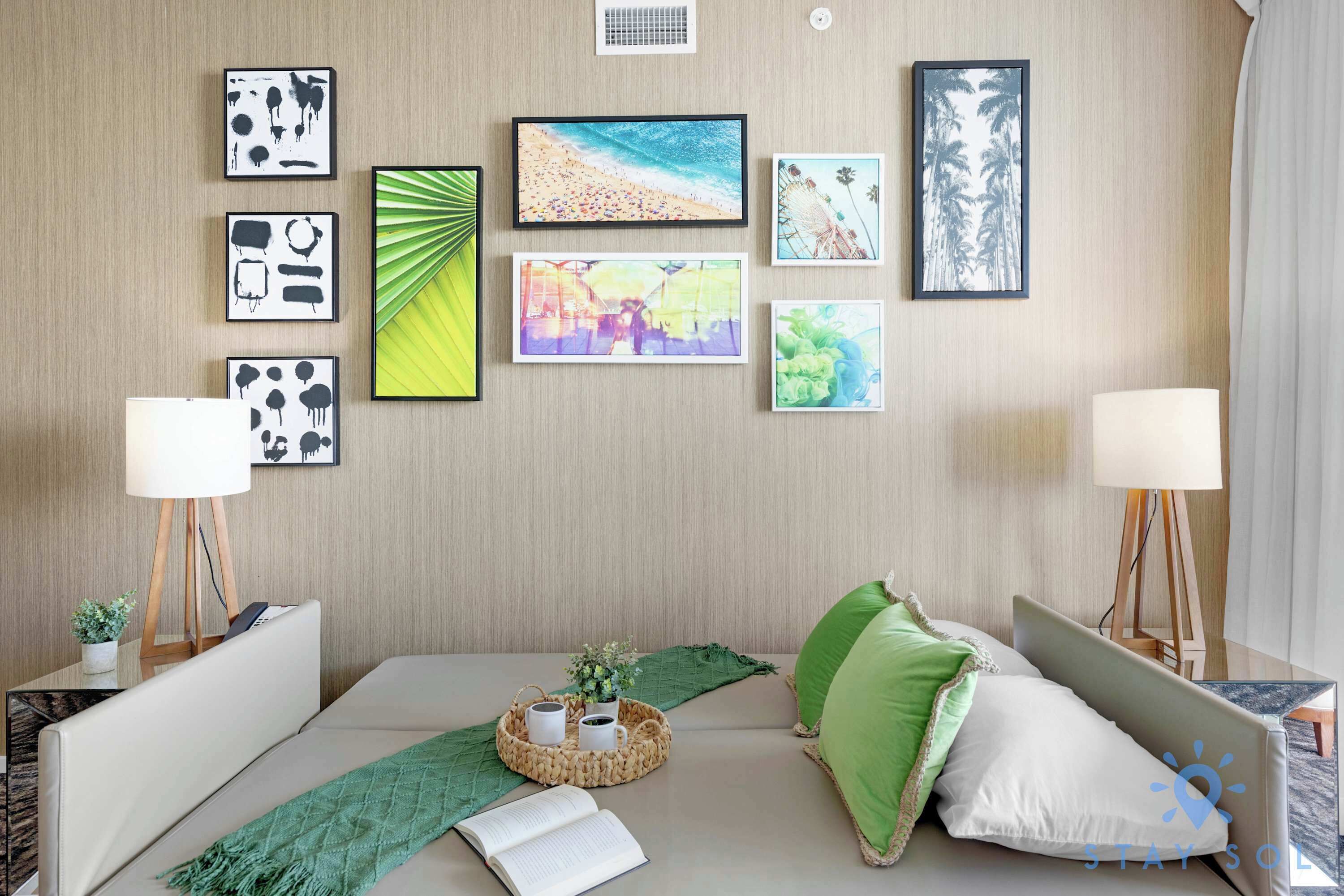 Habitación de hotel con cama, mesita de noche con lámpara, pared decorada con cuadros y ventana con cortinas.