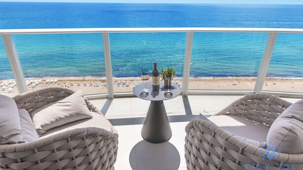 Varanda com móveis de vime, uma mesa com garrafa e copos, e vista para o mar e a praia.