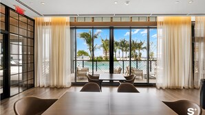 Un comedor moderno con una mesa grande y sillas, ventanas amplias con cortinas y vistas a palmeras y una piscina.