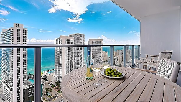 Een balkon met een houten tafel, twee wijnglazen, een fles wijn en een schaal druiven. Het balkon heeft uitzicht over een stadsgezicht met hoge gebouwen en een zeezicht.