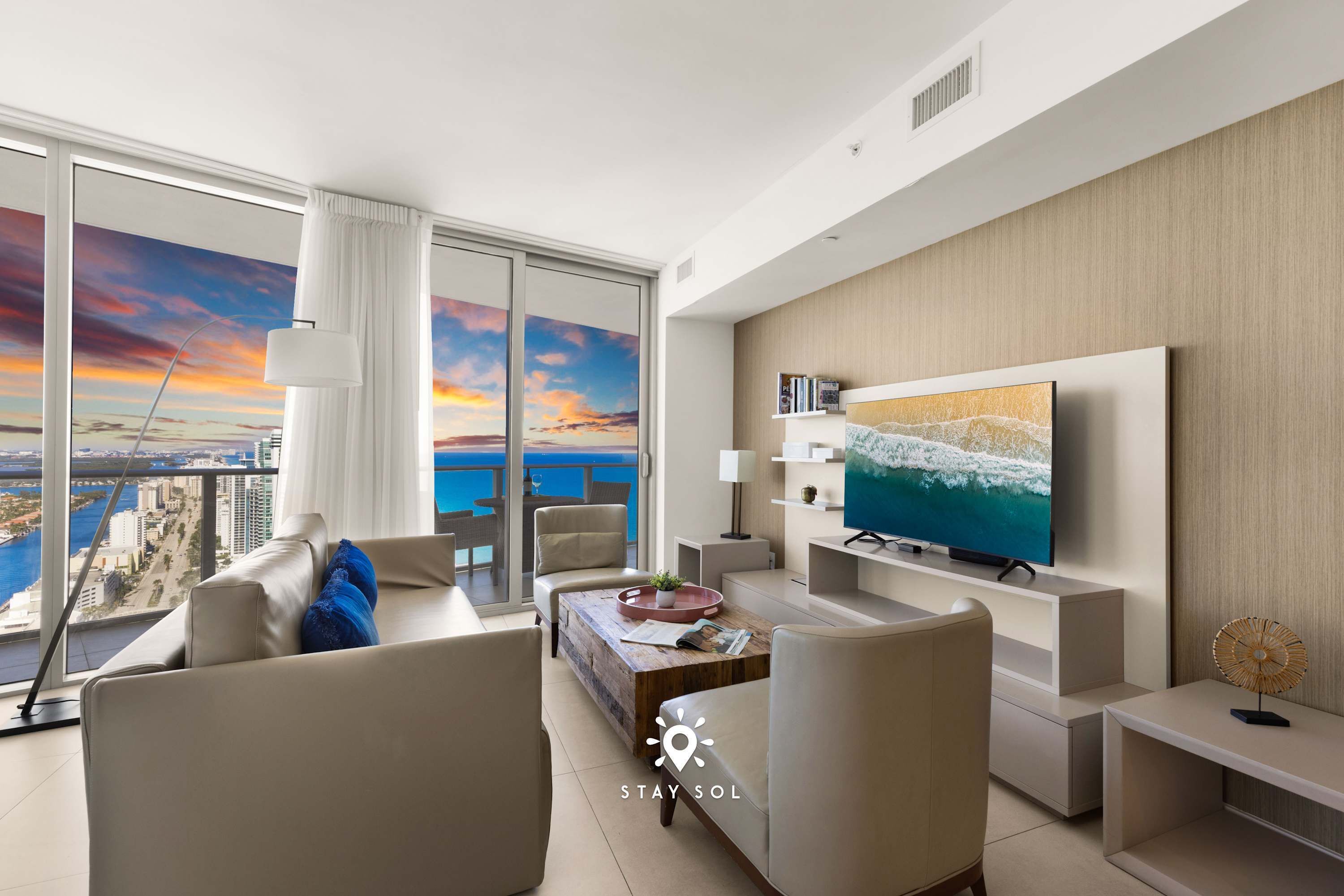 Una sala de estar moderna con televisor grande, asientos cómodos y vista a la ciudad y al océano al atardecer.