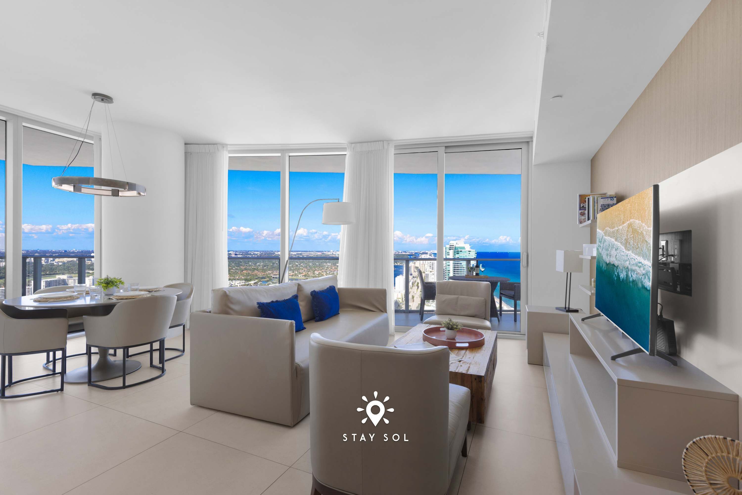 A modern living room with a dining area, large windows with a city view, and a flat-screen TV on the wall.