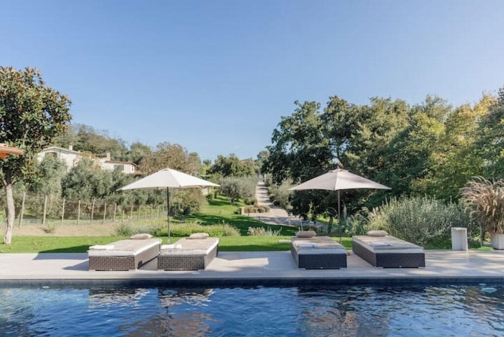 A poolside area with lounge chairs and umbrellas, surrounded by greenery and a clear sky.