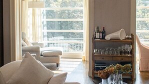 Una habitación con un sofá, una silla, un estante de madera con vasos y botellas, y un ventanal con vistas a árboles.
