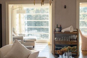 A room with a sofa, a chair, a wooden shelf with glasses and bottles, and a large window with a view of trees. - Villa Gatta Bianca - Villa with Pool in Natural Park near Rome (Formello)