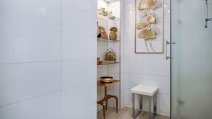 Une salle de bain moderne avec des murs recouverts de carreaux blancs, une douche en verre, un petit tabouret en bois, un banc blanc et des œuvres d’art décoratives sur les murs.