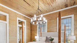 A bedroom with a wooden ceiling, a large bed with a headboard, a grey sofa, and a chandelier. - As Seen On "Restoring Galveston": Rustic Luxury (Galveston)