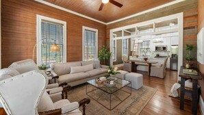 A spacious living room with a wooden ceiling, a sofa, armchairs, a glass coffee table, and a kitchen in the background. - As Seen On "Restoring Galveston": Rustic Luxury (Galveston)
