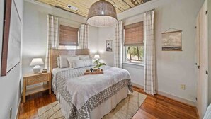 A bedroom with a wooden ceiling, a large bed with a patterned bedspread, two bedside lamps, and a hanging light fixture. - As Seen on "Restoring Galveston": Surf Shack (Galveston)