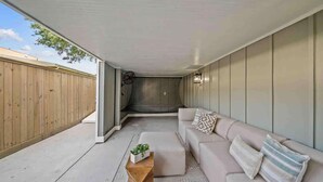 A covered patio area with a beige sofa, a wooden coffee table, and a small plant. - As Seen on "Restoring Galveston": Surf Shack (Galveston)