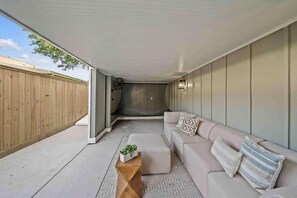 A covered patio area with a beige sofa, a wooden coffee table, and a small plant. - As Seen on "Restoring Galveston": Surf Shack (Galveston)