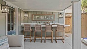 An outdoor bar area with a sign reading "EST. 3010 RUE. N", wicker chairs, and a wooden fence in the background. - As Seen on "Restoring Galveston": Surf Shack (Galveston)