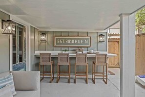 An outdoor bar area with a sign reading "EST. 3010 RUE. N", wicker chairs, and a wooden fence in the background. - As Seen on "Restoring Galveston": Surf Shack (Galveston)