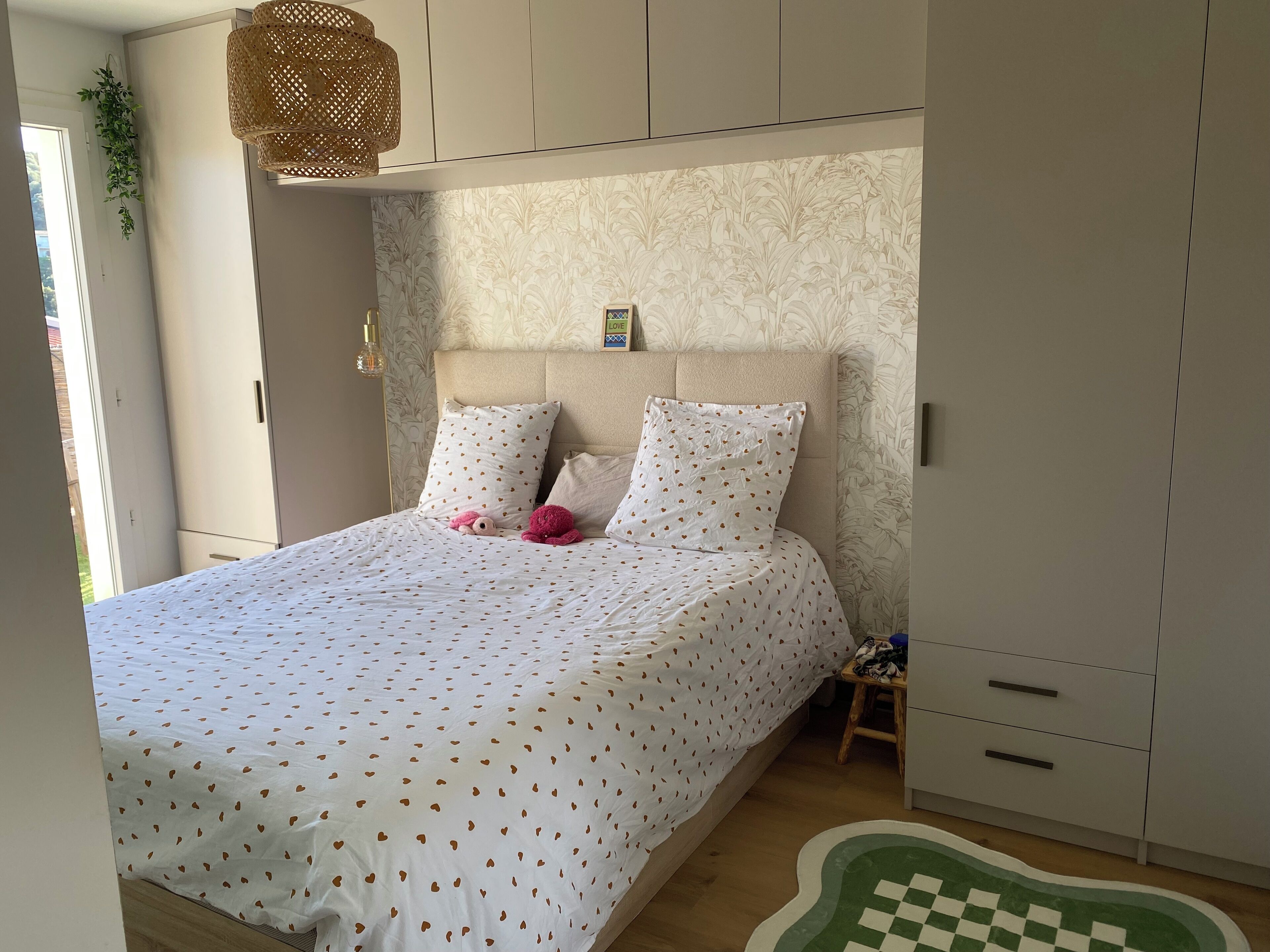 A bedroom with a bed, two pillows, a bedside table, a hanging light fixture, and a green and white rug.