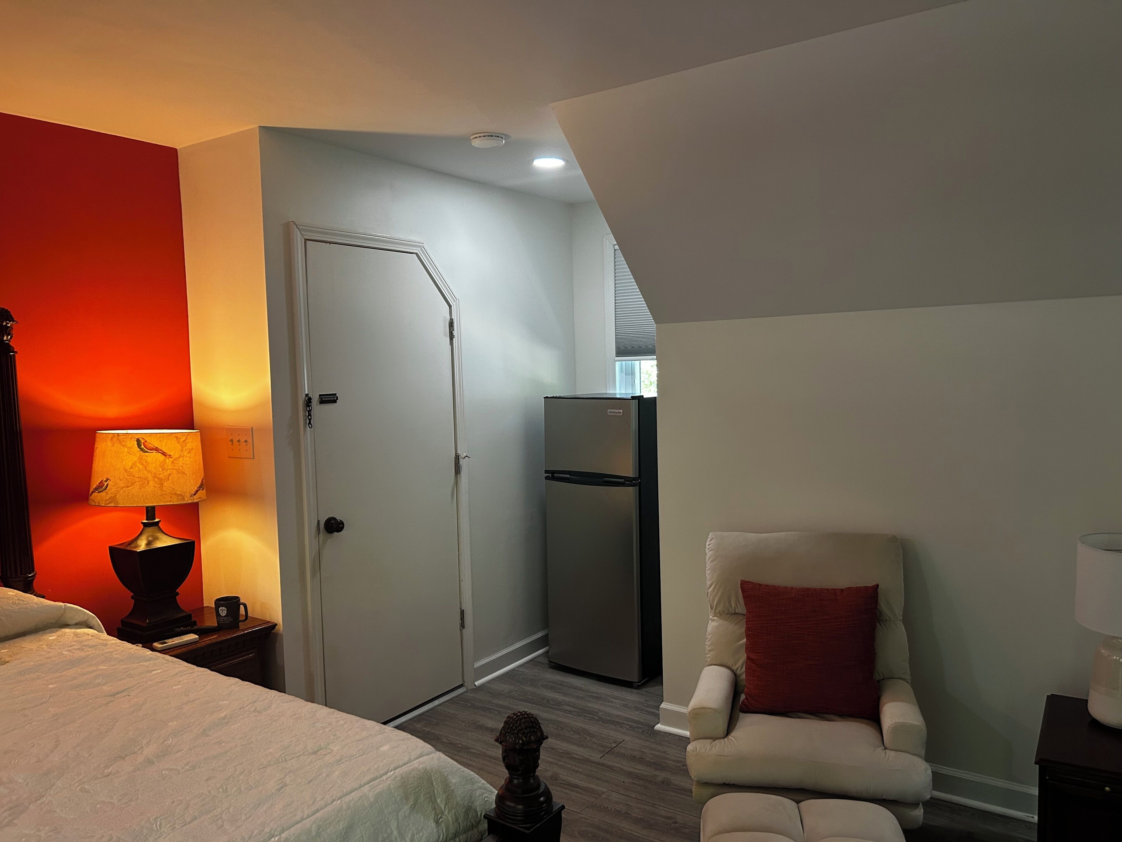 A hotel room with a bed, a chair, a lamp, a refrigerator, and a door.