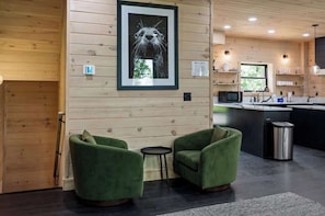 Een modern interieur met houten lambrisering, een ingelijste afbeelding, groene gestoffeerde stoelen, een kleine ronde tafel, een keukenhoek met een magnetron en een afvalbak.
