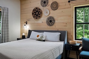 Een slaapkamer met een bed, nachtkastje, lamp en raam met uitzicht op bomen.