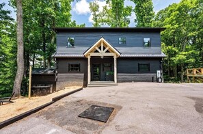 A two-story building with a dark exterior, a front porch with a triangular roof, and a small outdoor seating area. - 5-Bedroom Cabin with Glow Pool, Arcade Loft & Hot Tub, Sleeps 12 (Sevierville)