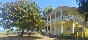 A yellow building with a balcony, surrounded by trees and a view of the sea.