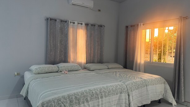A bedroom with a bed, curtains, an air conditioning unit, and a window with a view. - Tropikal Apartments B (Aldea de Perú)