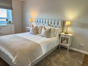 A neatly arranged bedroom with a large bed, two bedside tables, a nightstand, and a window with blinds. - The Kintyre - stylish beach apartment (Ayr)