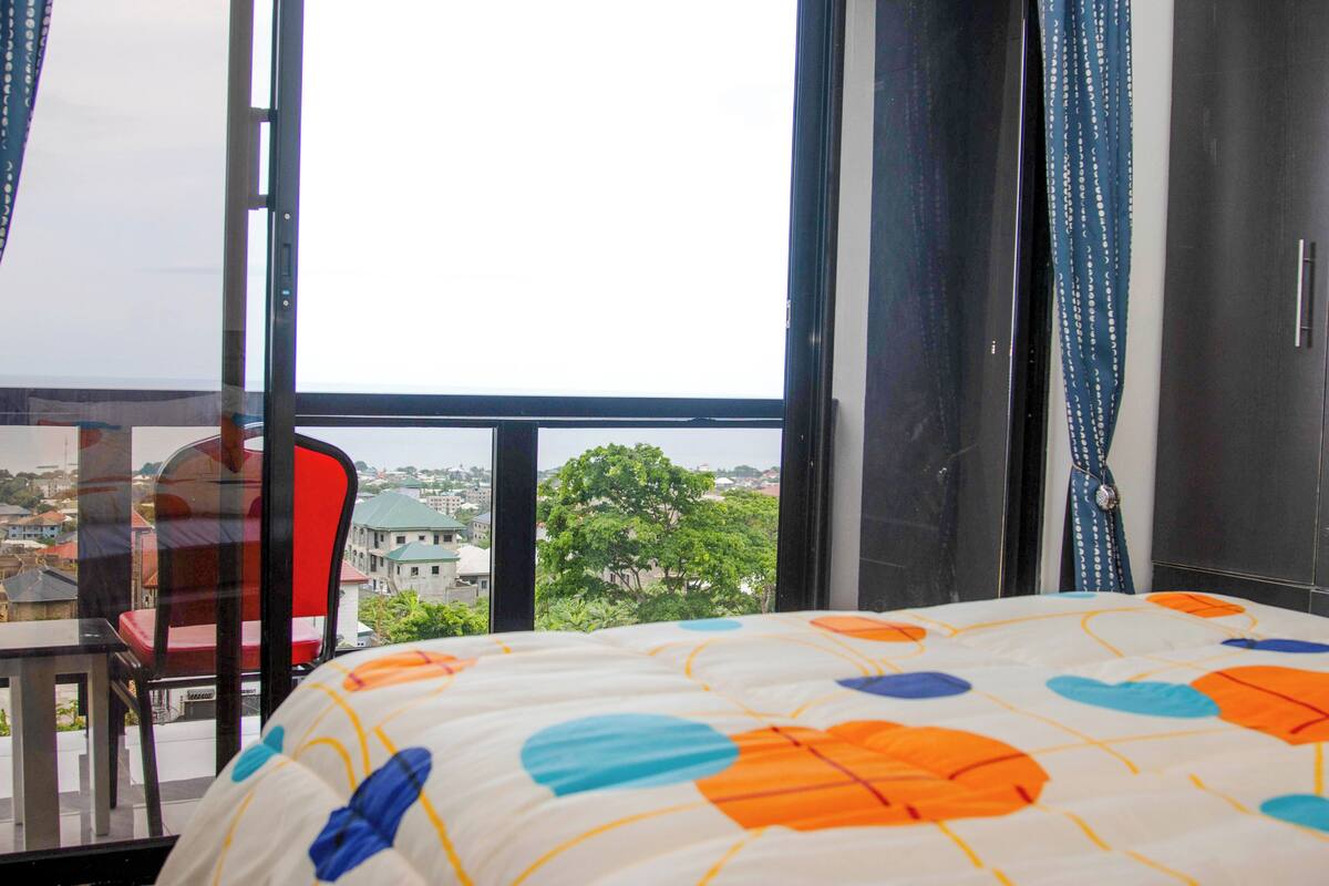 A hotel room with a bed, a chair, a table, and a large window offering a city view.