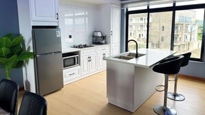 A modern kitchen with white cabinets, a stainless steel refrigerator, a microwave, a gas stove, and a large window offering a view of buildings. - K&K Luxurious Landing (Limbe)