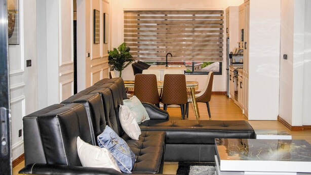 A modern living room with a black leather sofa, a dining area with a table and chairs, and a kitchen in the background. - K&K Luxurious Landing (Limbe)