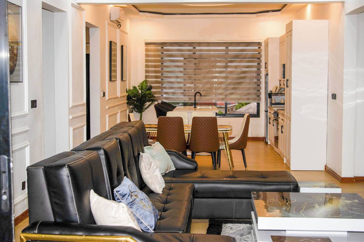 A modern living room with a black leather sofa, a dining area with a table and chairs, and a kitchen in the background.